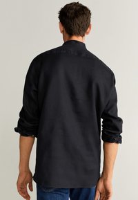 Uomo con le maniche rimboccate, indossante una camicia nera a maniche lunghe e jeans blu, in piedi e rivolto di spalle contro uno sfondo neutro.