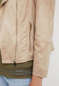 Béžová bunda z umělé semiše se zipem vpředu a boční kapsou. Vyznačuje se měkkou texturou a mírným leskem, s přiléhavým střihem.