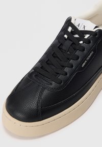 Zwarte leren sneaker met platte zwarte veters en een beige rubberen zool. Heeft een gestructureerde afwerking en subtiele branding aan de zijkant.