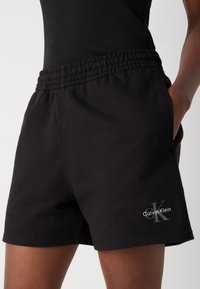Svarta bomullsshorts med elastisk midja, sidofickor och en liten grå "Calvin Klein"-logotyp på vänster ben. Enkel, avslappnad design.
