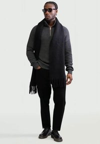 Bărbat purtând ochelari de soare negri, pulover gri închis cu țesătură cu zimți și fermoar, fular lung negru, pantaloni negri și pantofi negri, stând cu o mână în buzunar.