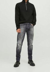 Svart zip-neck-tröja, grå slitna jeans och svarta snörkängor. Tröjan har ribbad mudd; jeansen har flera hål och en urblekt yta.