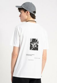 Młody mężczyzna w białej koszulce z czarną abstrakcyjną grafiką i tekstem na plecach, szarej czapce, czarnym naszyjniku, ustawiony bokiem na białym tle.