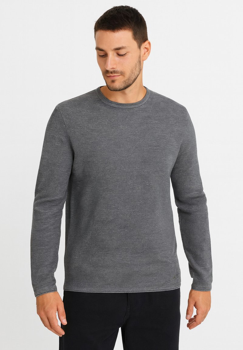 Graues Langarmshirt mit horizontalen Streifen, rundem Ausschnitt und glatter Textur. Über schwarzen Hosen getragen, steht das Modell vor einem neutralen Hintergrund.