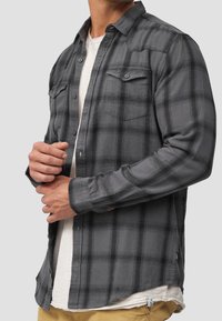 Chemise en flanelle à carreaux gris avec deux poches poitrine, boutonnage sur le devant et manches longues. La texture semble douce et légèrement brossée.