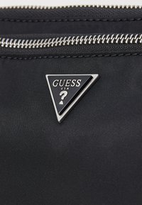 Bolsa de nailon negra con un cierre plateado. Un logo triangular en negro y plata de "GUESS" se muestra de manera prominente. Tela con textura visible.