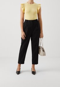 Kobieta w żółtej bluzce z falbanami i krótkim rękawem, czarnych, wysokich spodniach 7/8, czarnych szpilkach, trzymająca beżową torebkę z marszczeniami.