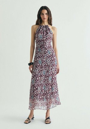 Mujer de pie con un vestido sin mangas hasta el tobillo con patrones geométricos en púrpura, blanco, azul y rojo y sandalias negras con tiras.