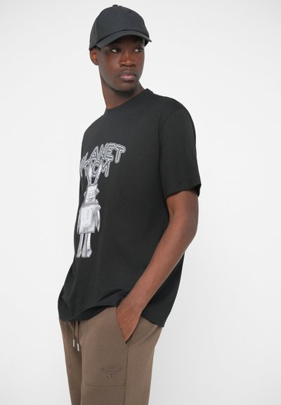 Muškarac u crnoj baseball kapi, crnoj majici s robotom i natpisom "PLANET COM", i smešnim smešnim hlačama, stoji s rukama u džepovima.