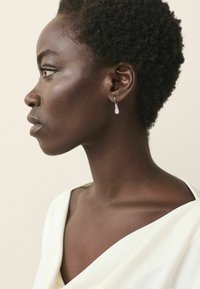 Hopea renkaallinen korvakoru, jossa on tipan muotoinen vaaleanlila yksityiskohta, esillä neutraalilla taustalla, joka korostaa korvakorun ainutlaatuista muotoilua.