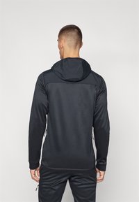 Giacca nera con cappuccio e maniche lunghe, caratterizzata da una texture liscia e un pannello superiore a contrasto. Include cerniere laterali e polsini elasticizzati.