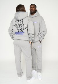 Graues Sweatshirt und Sweatpants-Set mit Zugbändern. Enthält ein großes schwarzes Grafikdesign auf dem Rücken und ein lila Markenlogo auf der Vorderseite.