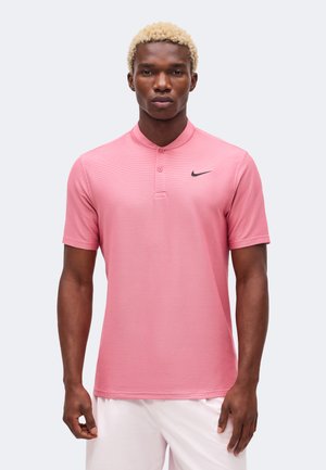 Mand med kort blondt hår iført en pink Nike kortærmet polo og hvide shorts, stående foran en ensfarvet baggrund.