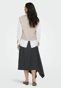 Beżowy sweter bez rękawów nałożony na białą koszulę z rozciętymi rękawami, w połączeniu z asymetryczną spódnicą w ciemne paski i brązowym obuwiem.