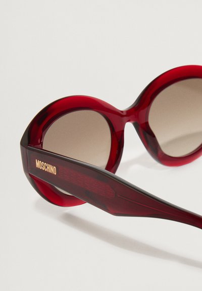 Punased ülemõõdus prillid sujuva plastraami, gradient pruunide läätsede ja tekstuuriga käe ning brändinime "Moschino" kullakirjas.