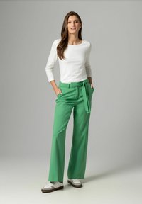 Grüne weite Hosen mit gebundenem Hüftgurt, kombiniert mit einem weißen Langarmoberteil. Merkmale sind ein glattes Material und eine lockere Passform. Weiße Sneakers vervollständigen den Look.