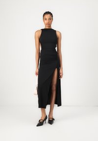 Melnā bez piedurknēm kleita ar sānu izgriezumu un sakirnājumiem, izgatavota no elastīga auduma, apvienota ar melnām augstpapēžu kurpēm un mazu rozā rokassomu.