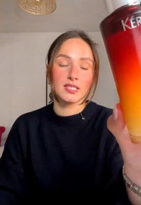 Giovane donna con gli occhi chiusi che tiene un bicchiere alto con una bevanda a gradiente dal colore arancione scuro a chiaro, in ambiente interno.