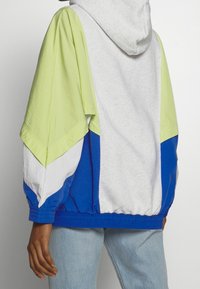 Giacca con cappuccio a blocchi di colore, caratterizzata da maniche corte, corpo grigio e accenti vivaci in verde, blu e bianco. Tessuto leggero con vita elastica.