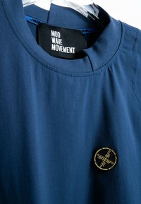 Camisa azul de cuello redondo con etiqueta negra que dice "MOVIMIENTO MOD WAVE" y un parche bordado redondo negro y dorado cerca del dobladillo.