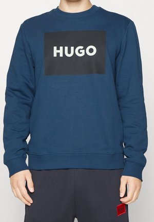 Person trägt ein blaues Sweatshirt mit einem schwarzen Quadrat und weißem "HUGO"-Schriftzug in der Brustmitte, kombiniert mit einer dunklen Hose mit rotem Aufnäher.