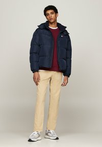 Giacca imbottita blu navy con colletto alto, sezioni imbottite e chiusura con zip, indossata sopra un felpa bordeaux e pantaloni beige con sneakers chiare.