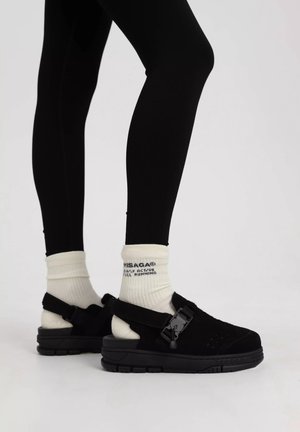 Zwarte schoenen met een gestructureerde bovenkant, verstelbare band en dikke zool. Gecombineerd met crèmekleurige sokken met zwarte tekst, gedragen met zwarte leggings.