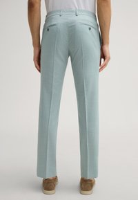 Helle blaue Hose mit einer glatten Textur, ausgestattet mit zwei Gesäßtaschen mit Knopfverschluss und einem maßgeschneiderten Schnitt, getragen mit braunen Schuhen.