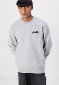 Gray sweatshirt with ribbed cuffs and hem, featuring a black logo on the left chest. Fabric appears soft and textured. Casual fit.