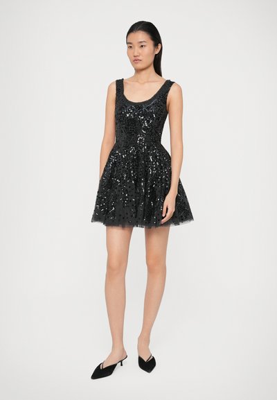 Žena v černých šatech bez rukávů posetých flitry a s rozšířenou sukní, v černých botách na podpatku s úzkou špičkou, stojící na bílém pozadí.