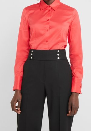 Persona che indossa una camicia color corallo brillante con maniche lunghe abbottonata infilata in pantaloni neri a vita alta con dettagli di bottoni argentati.