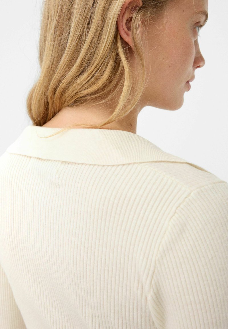 Maglione a coste di color crema con colletto rotondo, che mostra texture e una leggera lucentezza. La parte posteriore del colletto presenta dettagli di cucitura sottili.