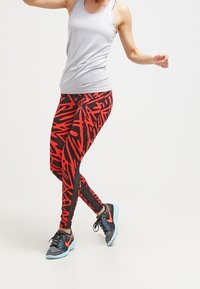 Canotta sportiva grigia, leggings a motivo rosso e nero e sneakers nere con accenti arancioni. Focalizzarsi sul design e la vestibilità dell'abbigliamento sportivo.