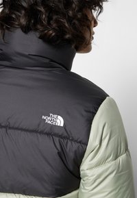 Czarna i jasnozielona kurtka puchowa z wysokim kołnierzem. Zawiera białe logo The North Face z tyłu, prezentujące pikowaną fakturę.