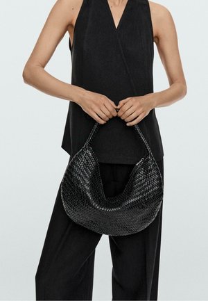 Frau in ärmellosem schwarzem Oberteil und Hose hält vor sich mit beiden Händen eine große schwarze geflochtene Ledertasche.