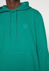 Green cotton hoodie featuring a front kangaroo pocket, adjustable drawstring hood, and a small embossed logo on the chest.