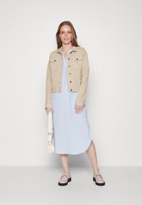 Beige denim takki vaaleansinisellä mekolla, jota korostavat pyöreä valkoinen laukku ja vaaleat loafertossut mustilla pohjilla. Sileä kangas ja rento malli.
