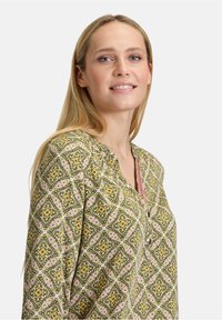 Zielona tunika w wzory z kwiatowymi motywami w odcieniach żółtego i różowego, z dekoltem w kształcie litery V, wykonaną z miękkiego materiału i z detalem guzika przy szyi.