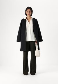 Black oversized coat, white shirt, textured olive green pants, and a light beige bag with a logo. Smooth fabrics and relaxed silhouette.