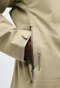 Χέρι σε τσέπη μπεζ αδιάβροχης ζακέτας με ορατό φερμουάρ και μανσέτα με βέλκρο, που δείχνει το λογότυπο της μάρκας Houdini.