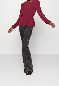 Person som bär en långärmad vinröd blus och högmidjade urtvättade svarta jeans med svarta kraftiga skor mot en neutral bakgrund.