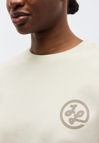 Gros plan sur une personne portant un t-shirt beige clair à col rond avec un logo circulaire arborant des lettres stylisées "J" et "P" sur la poitrine.