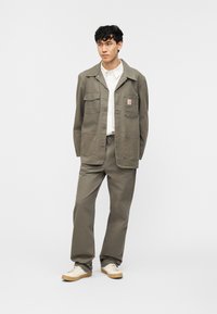 Νεαρός άνδρας που στέκεται φορώντας γκρι επαγγελματικό μπουφάν και παντελόνι, λευκό πουκάμισο και μπεζ αθλητικά παπούτσια σε ανοιχτό φόντο.