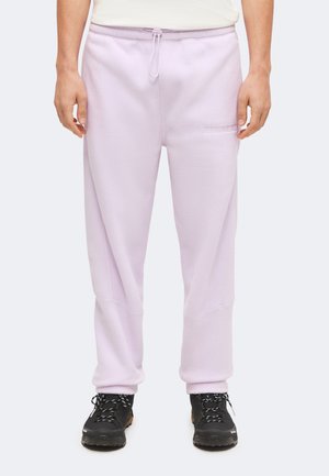Homme portant un jogging en polaire violet clair avec une taille à cordon et des chaussures de randonnée noires, debout devant un fond uni.