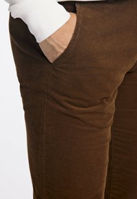 Main dans la poche avant d'un pantalon en velours côtelé marron, portée avec une manche de chemise blanche à manches longues visible.