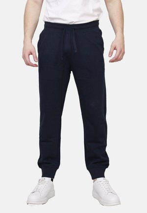 Uomo che indossa pantaloni jogger neri con coulisse e sneakers bianche, in piedi davanti a uno sfondo chiaro e uniforme.