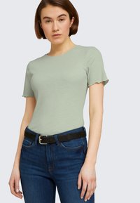 Svetlozelené rebrované tričko s krátkym rukávom a zúbkovanými okrajmi, skombinované s tmavomodrými džínsami a čiernym opaskom so striebornou sponou.