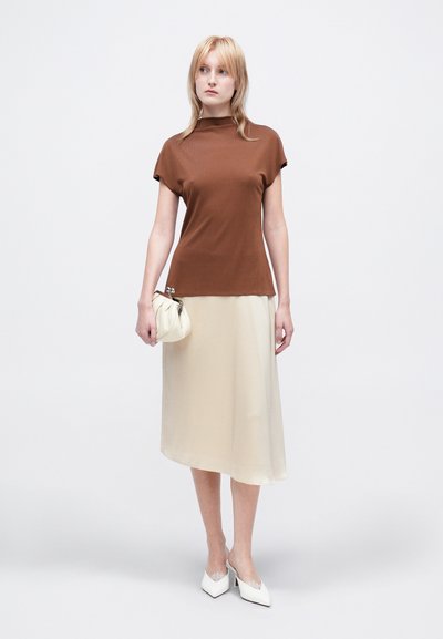 Top scurt de culoare maro cu guler înalt, asortat cu o fustă midi din satin crem. Modelul ține o geantă adunată de culoare deschisă. Pantofi cu toc alba cu accente.