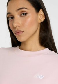 Sudadera de color rosa claro con cuello redondo y ribete en el cuello. Presenta un sutil logo blanco en el pecho. Textura de tela suave.