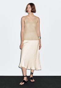 Nøgenfarvet striktop og cremefarvet satinskjørt; toppen har en rund halsudskæring og tekstureret mønster, skørtet har en flydende kant. Sorte flade sandaler fuldender looket.